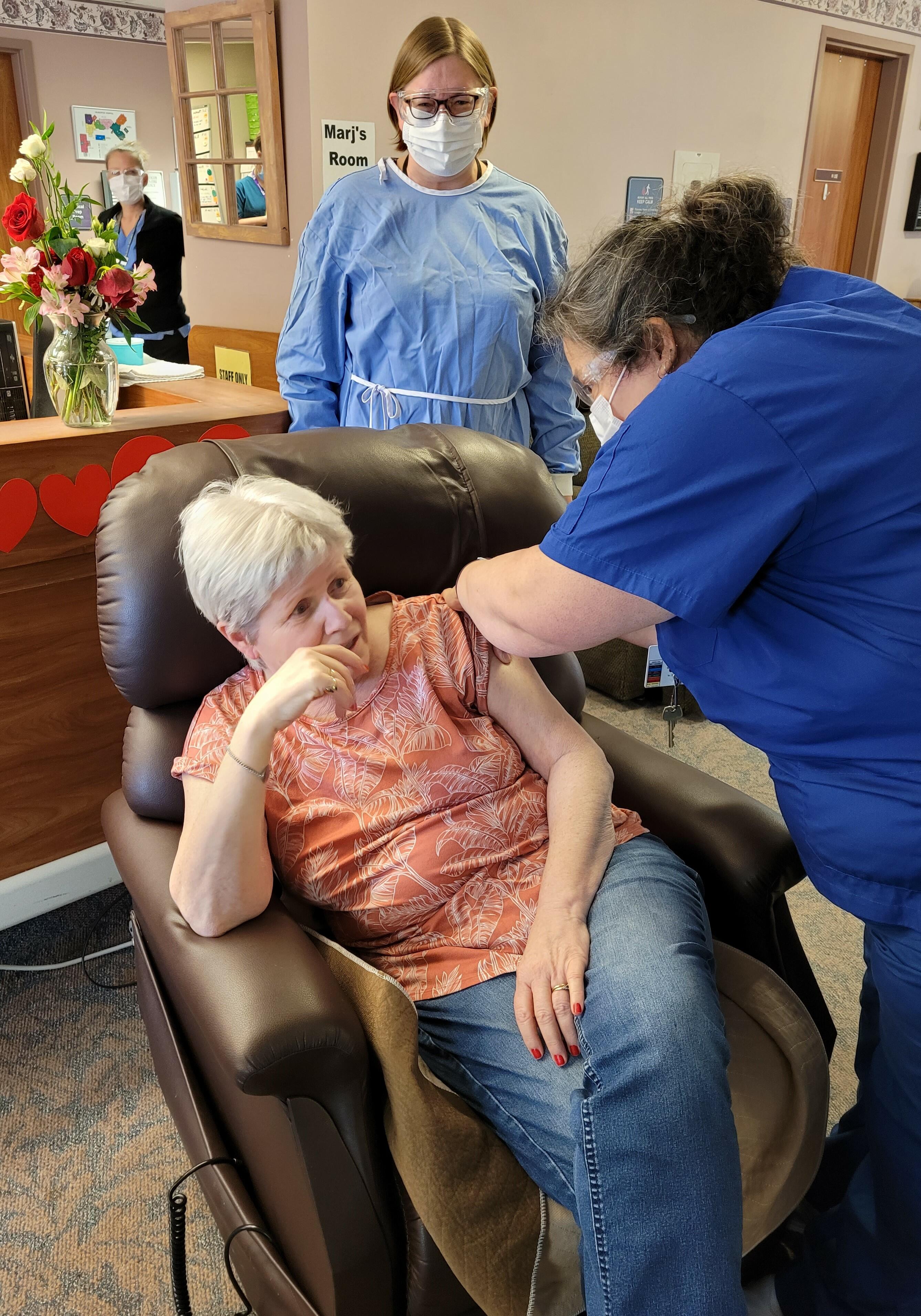 Nancy McElwain, the first Highland Wood Long-Term Care resident to receive the Moderna COVID-19 vaccine on February 8, 2021. Also pictured are a Sue Long, Highland Wood Nurse (in dark blue) administering the vaccination, and HHHS President & CEO Carolyn Plummer (in light blue), looking on. Angela Brannigan, a nurse at Highland Wood looks on in the background.